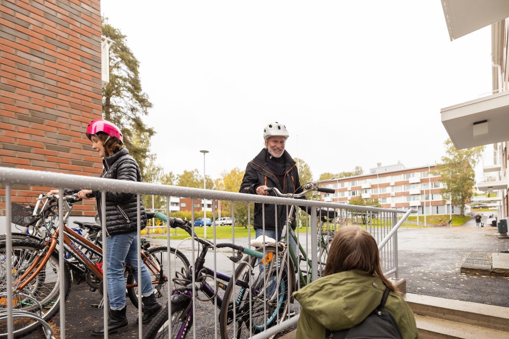 Nainen kävelee pihaportaita ylöspäin ja näkee ylätasanteella kaksi naapuriaan, jotka ovat lähdössä pyöräilemään. Naapureilla on päässään pyöräilykypärät.